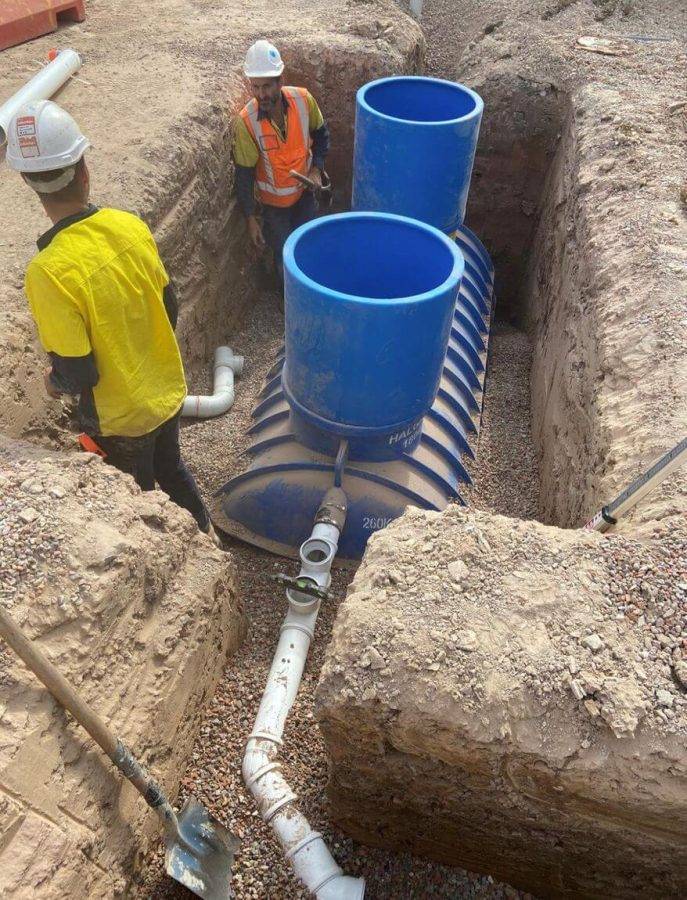 Construction workers in safety gear installing plumbing with blue tanks and an excavated trench.