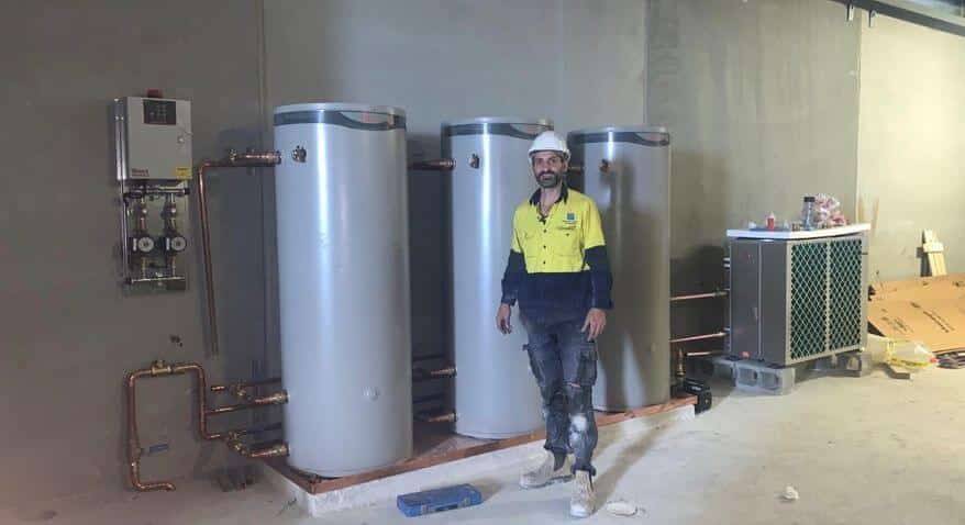 Man in hard hat at industrial plumbing site with silver tanks and copper piping by Priority Plus Plumbing.
