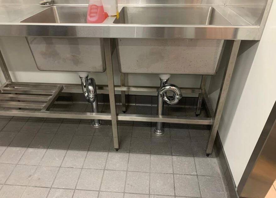 Stainless steel sinks in a commercial kitchen by Priority Plus Plumbing, featuring fixtures and drainage pipes.